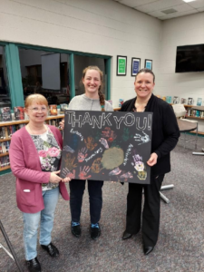 Cheryl Davis and Samantha Mitchell holding a large, black poster with the words "Thank You" and painted with hands and foot prints and the words "Explorers" "Innovators" and "Pathfinders". Jennifer McKittrick is also holding it, receiving it from CDA. 
